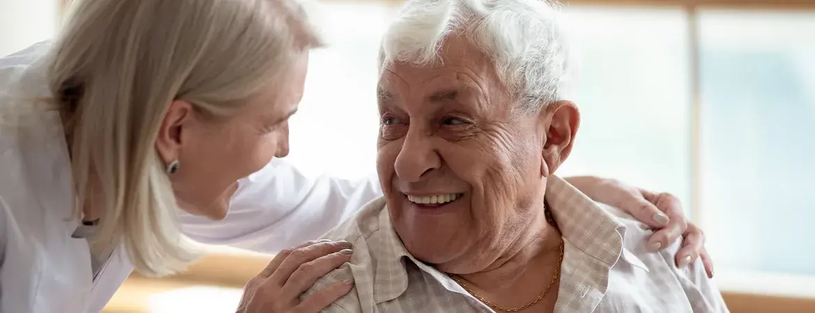 A woman has placed both of her hands on the shoulder of an elderly man and both are looking at each other and smiling