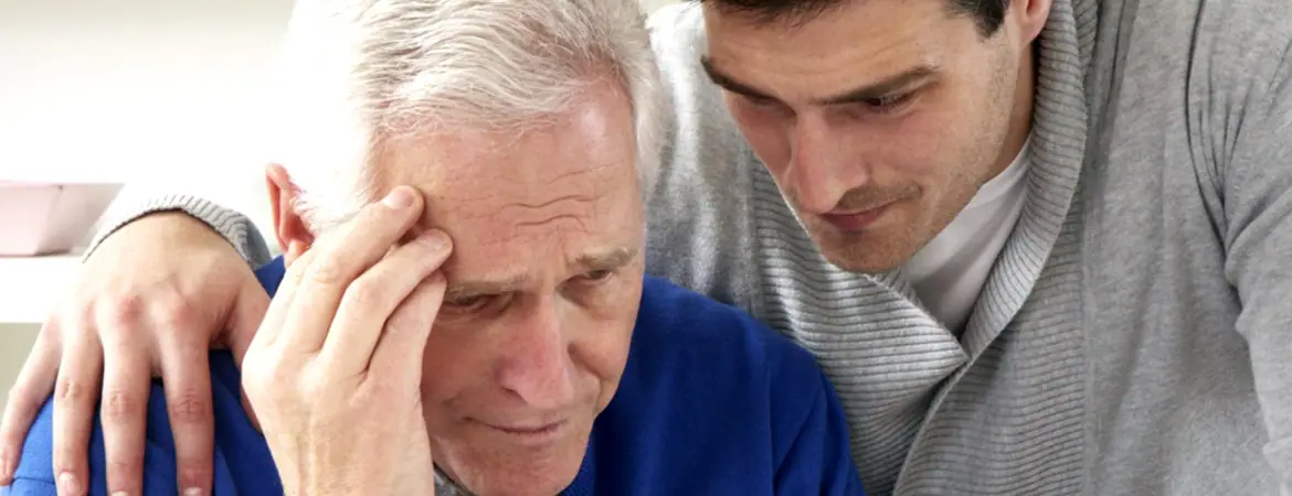 An elderly person holding his head with one hand is being consoled by a young man